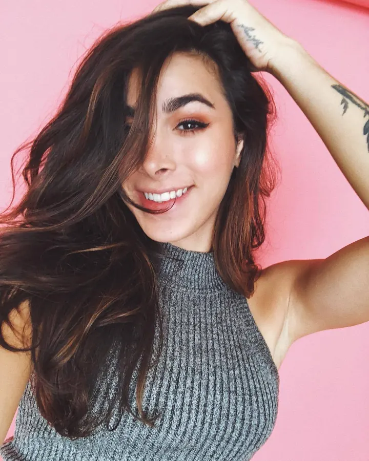 Valentine en pull gris sans manches, fond rose, sourire, main dans les cheveux