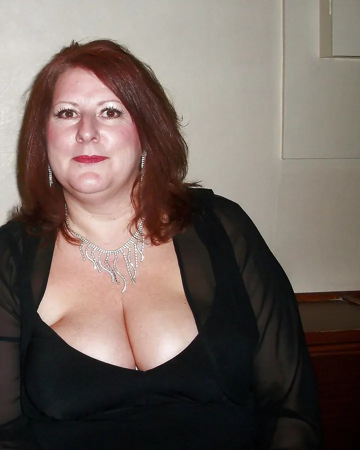 Femme aux cheveux roux, vêtue d'une robe noire décolletée, portant un collier argenté et des boucles d'oreilles, regardant l'appareil photo.