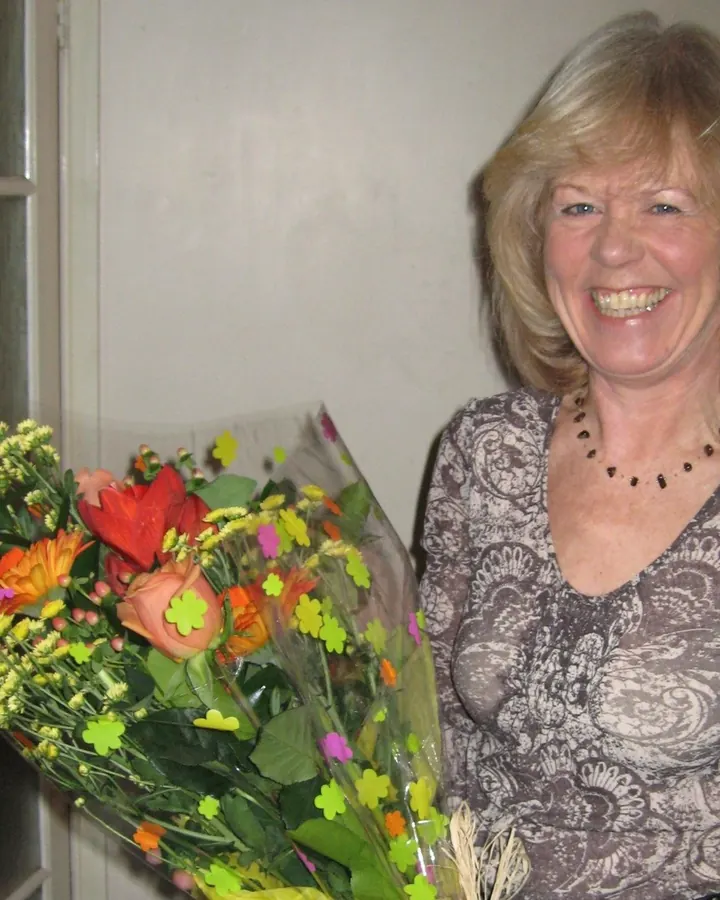 Femme aux cheveux blonds clairs, souriante largement, tenant un grand bouquet de fleurs colorées.