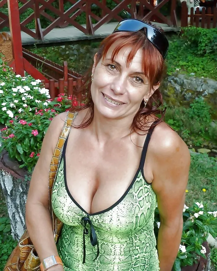 Femme aux cheveux roux en haut vert serpenté, debout dans un jardin fleuri, regardant l'appareil avec un sourire chaleureux.