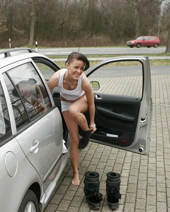 Femme aux cheveux foncés, souriante, sortant d’une voiture blanche, portant un haut blanc et un short court, pieds nus, chaussures noires à côté d’elle.