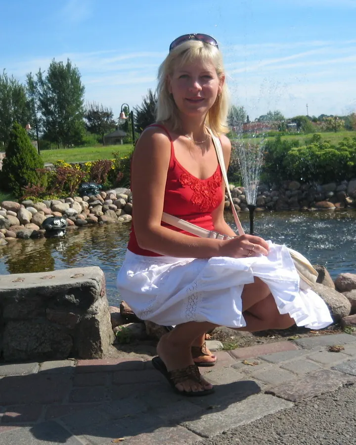 Femme blonde en robe rouge et jupe blanche, assise sur un bord de piscine, souriante, avec un petit jet d'eau derrière elle.