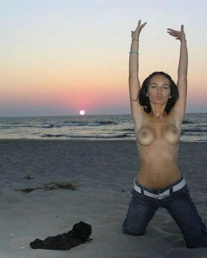 Femme nue, genoux fléchis sur le sable, bras levés, portant un jean, devant une mer au coucher du soleil.