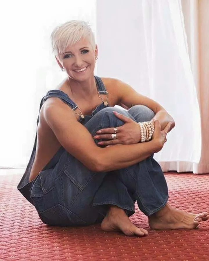 Femme aux cheveux blancs assise sur un tapis rouge en salopette en jean, souriante